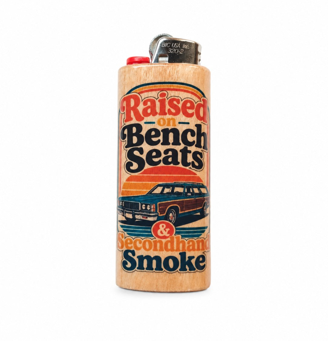 A novelty lighter with a light wood grain finish, featuring a retro-style printed graphic that reads 'Raised on Bench Seats & Secondhand Smoke' with an illustration of a station wagon. It has a red ignition button and a silver metal top.